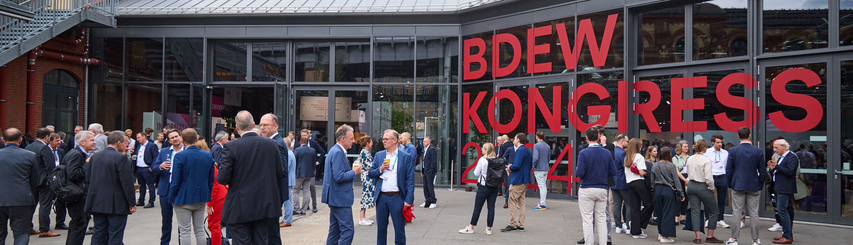 Menschen unterhalten sich vor einer mit dem Logo des BDEW Kongress 2024 beklebten Glasfassade