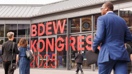 Menschen laufen auf eine Glasfassade mit der Aufschrift 'BDEW Kongress 2025' zu